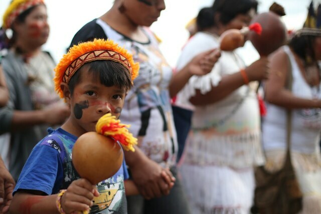 Crian&ccedil;a com pintura no rosto segura um chocalho de caba&ccedil;a, enquanto outras pessoas carregam instrumentos semelhantes ao fundo.