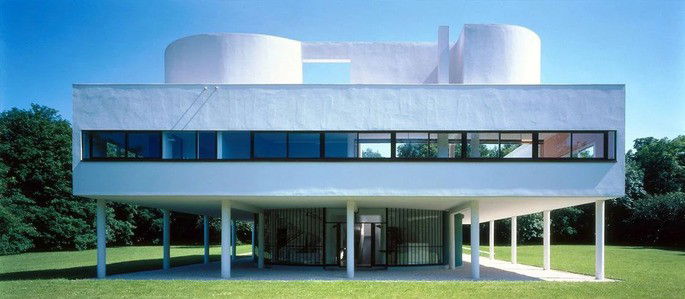 Villa Savoye. Residência construída pelo arquiteto Le Corbusier entre 1928 e 1931. Trata-se de fotografia da fachada de uma casa branca, construída sobre pilares, também brancos, que sustentam o imóvel um andar acima do nível do chão. A casa é bastante geométrica, retangular, e conta com amplas vidraças na fachada, permitindo que muita luz solar a ilumine.