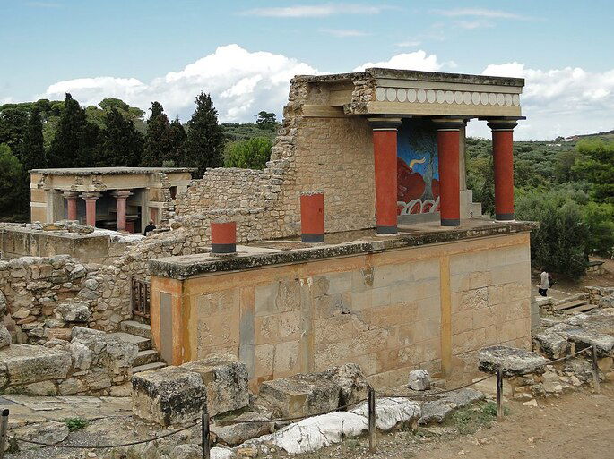 Palácio de Knossos, em Creta. È possível ver um palácio parcialmente em ruínas, com colunas vermelhas e afrescos nas paredes.