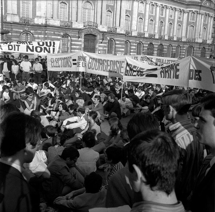 Manifestantes organizam um protesto na França em 1968. Há manifestantes sentados na rua, cartazes e bandeiras com slogans.