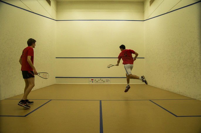 Dois jogadores de Squash em uma quadra fechada, com as marcações necessárias para a modalidade. O homem da direita está realizando a rebatida com a sua raquete, enquanto o homem da esquerda espera para tentar realizar a sua jogada.